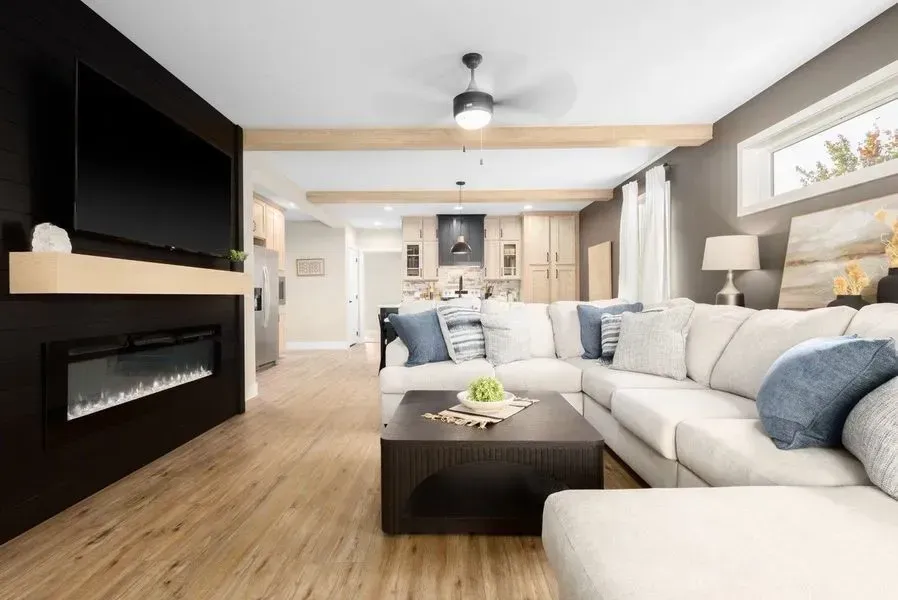 A modern living room with a black fireplace wall, light wood floors, a beige sectional sofa, and a kitchen in the background.