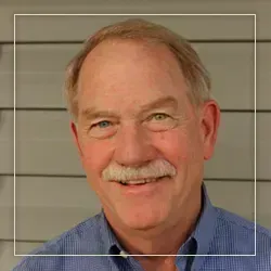 A smiling person with a mustache, wearing a blue button-down shirt, positioned in front of a gray wall.