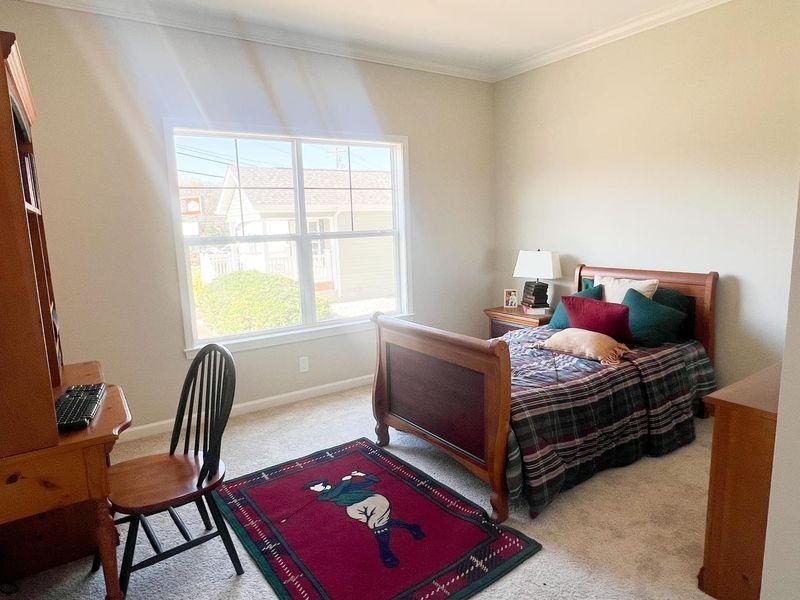 A bedroom with a wooden bed, a desk and chair, a small red rug, and a large window in a neutrally painted room.
