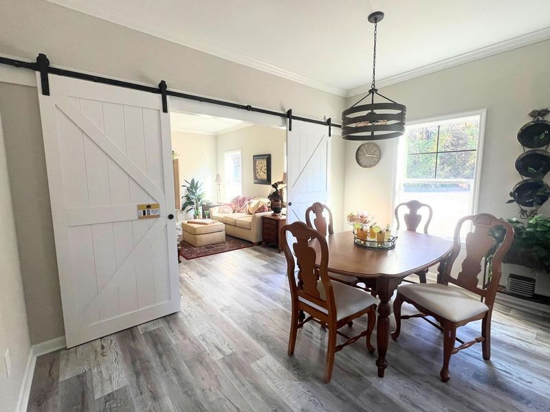 A dining room with a wooden table and chairs, illuminated by a pendant light, adjacent to a living area with barn doors.