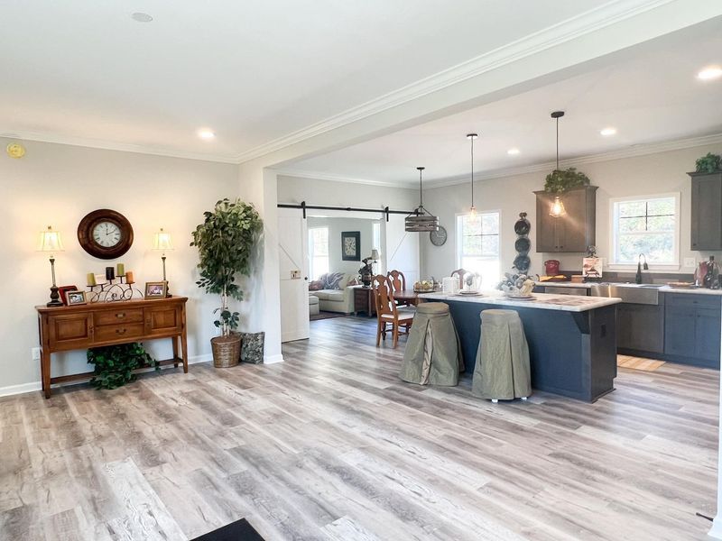 A bright, open-plan kitchen and living space with grey wood-look floors, a dark island, and a wooden console table.