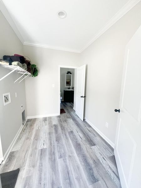 A laundry room with light gray wood-look flooring, white walls, wire shelving, and an open door leading to a bathroom.