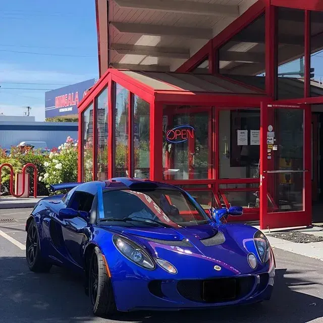 A vibrant blue Lotus sports car parked in front of a building with a red-framed glass entrance and an 