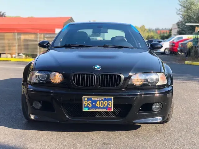 A black BMW E46 M3 coupe parked outdoors, facing forward with its headlights on and an Oregon license plate.