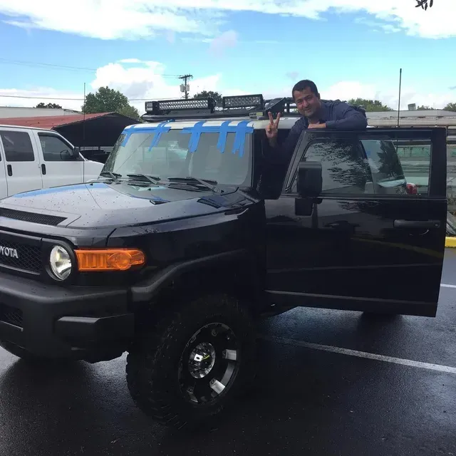 A person making a peace sign while leaning out of the driver's side door of a black Toyota FJ Cruiser in a parking lot.