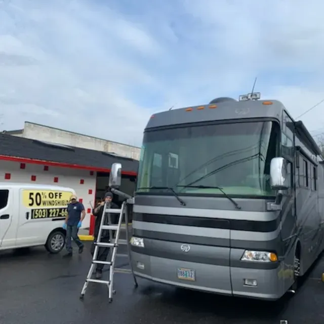 A technician on a ladder works on the windshield of a large grey RV, with a service van parked nearby in a paved lot.