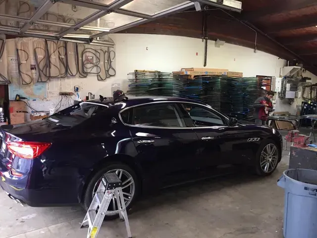 A dark blue Maserati sedan sits in an auto repair shop with a small ladder and a large blue trash bin in the foreground.
