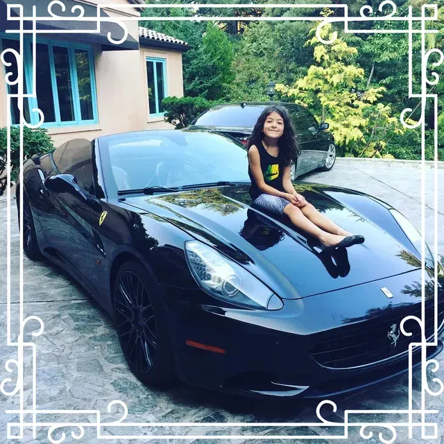 A young person sits on the hood of a black Ferrari parked in a driveway in front of a house.