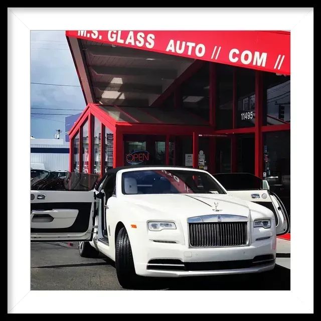 A white Rolls-Royce with its doors open parked in front of an auto glass shop with a red exterior.