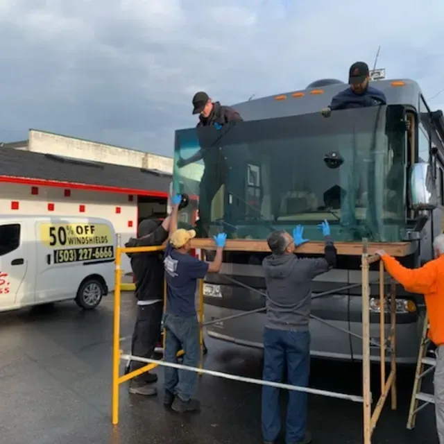 Five people work together to install a large windshield into a motorhome using a yellow scaffolding frame.