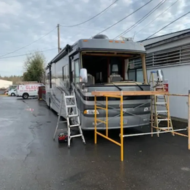 A grey motorhome parked outside with its windshield removed, surrounded by ladders and scaffolding for repairs.