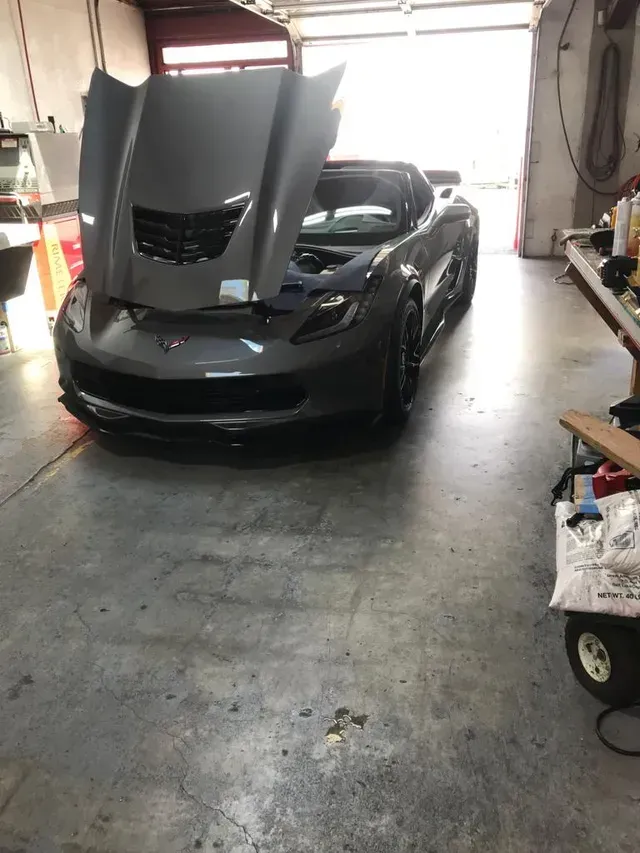 A gray Chevrolet Corvette with its hood raised in a brightly lit automotive repair garage.