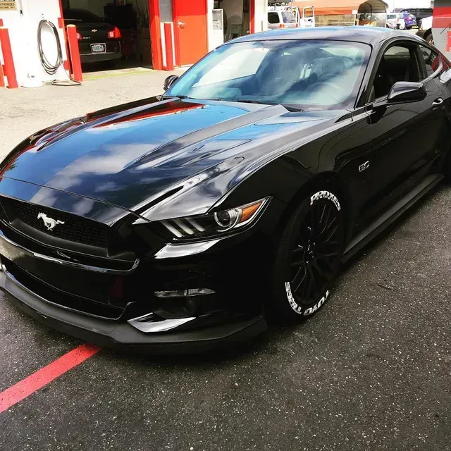 A shiny black Ford Mustang parked outdoors, featuring a prominent front splitter and custom white-lettered tires.