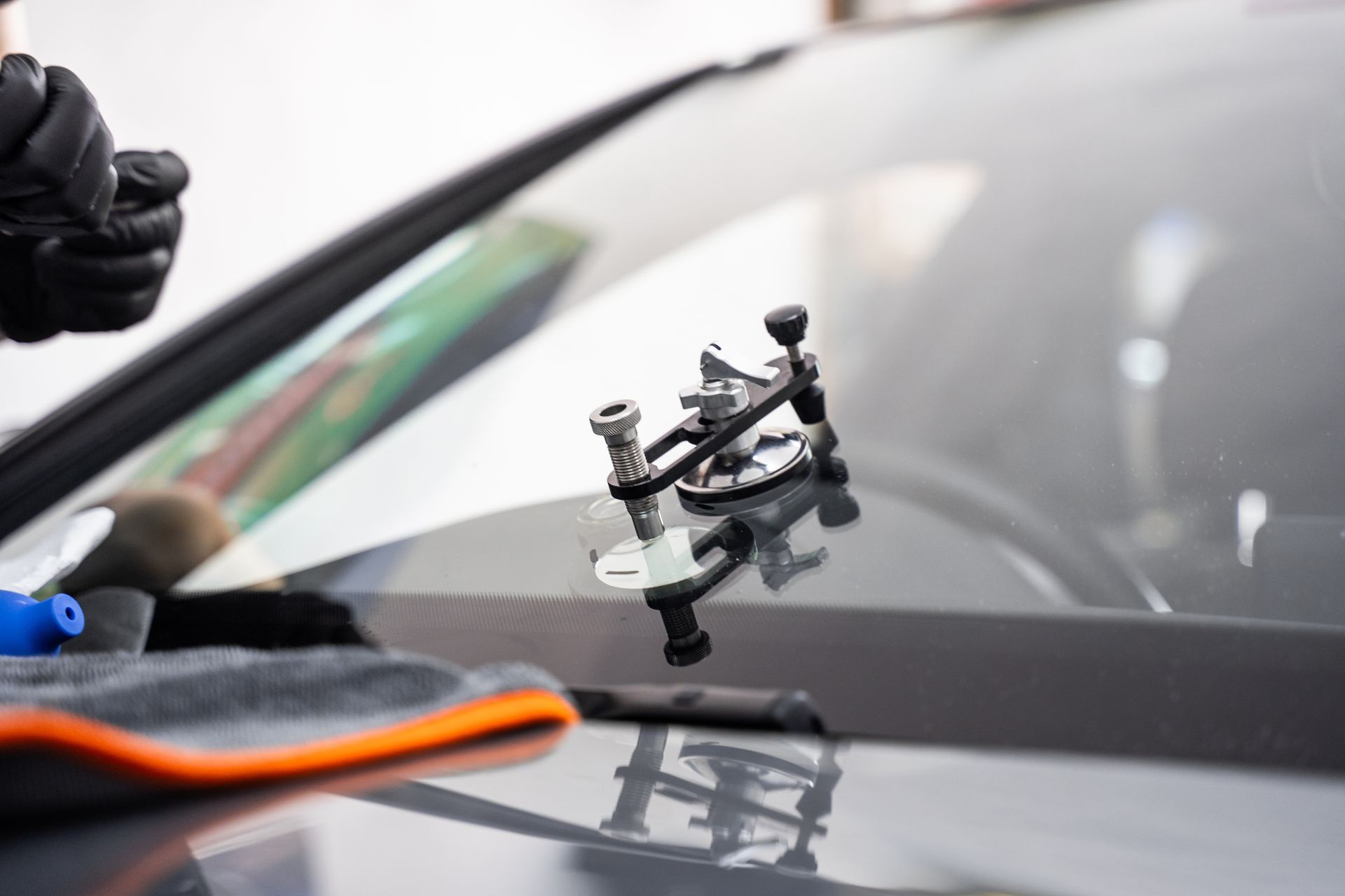 A tool for repairing a windshield crack is attached to a car's glass, with a person wearing black gloves in the background.