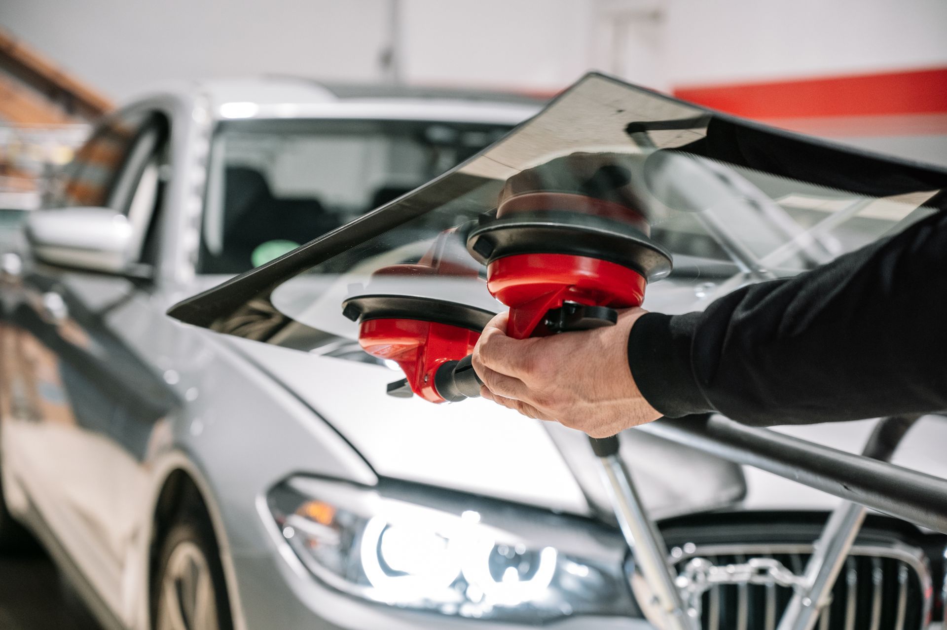 A person uses red suction tools to hold a new car windshield in front of a silver BMW in a workshop.