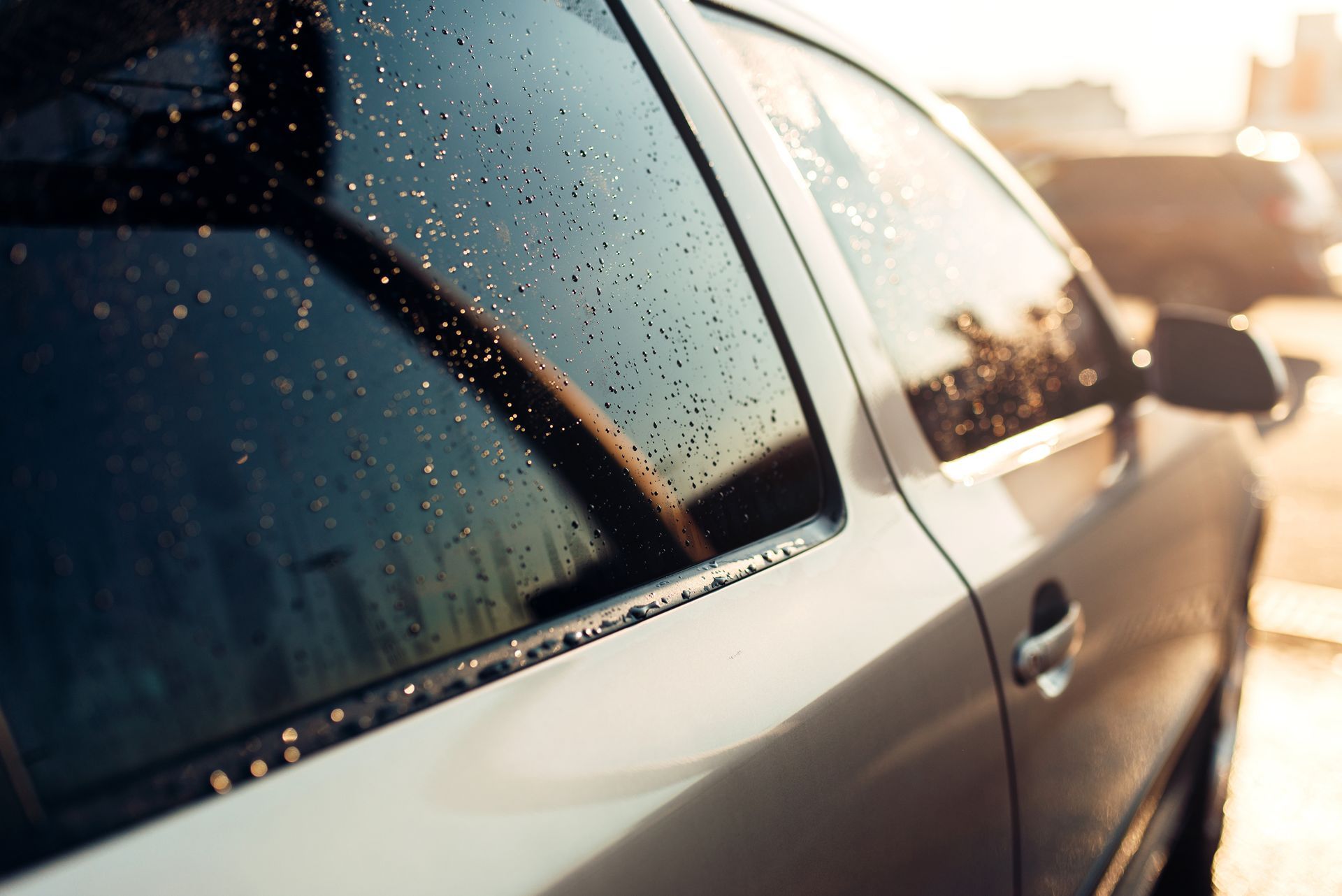 Silver car window with water droplets, reflecting sunlight in a close-up shot.