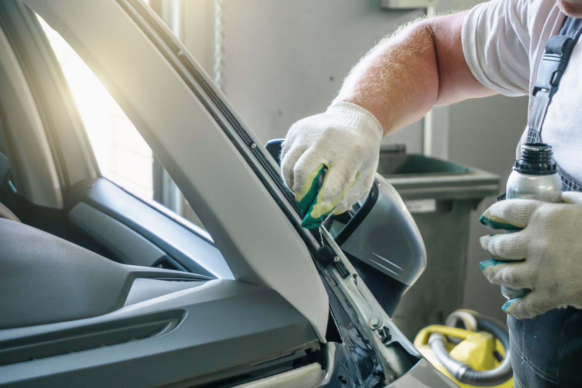 Person wearing gloves applying liquid to a car's side mirror in a workshop.