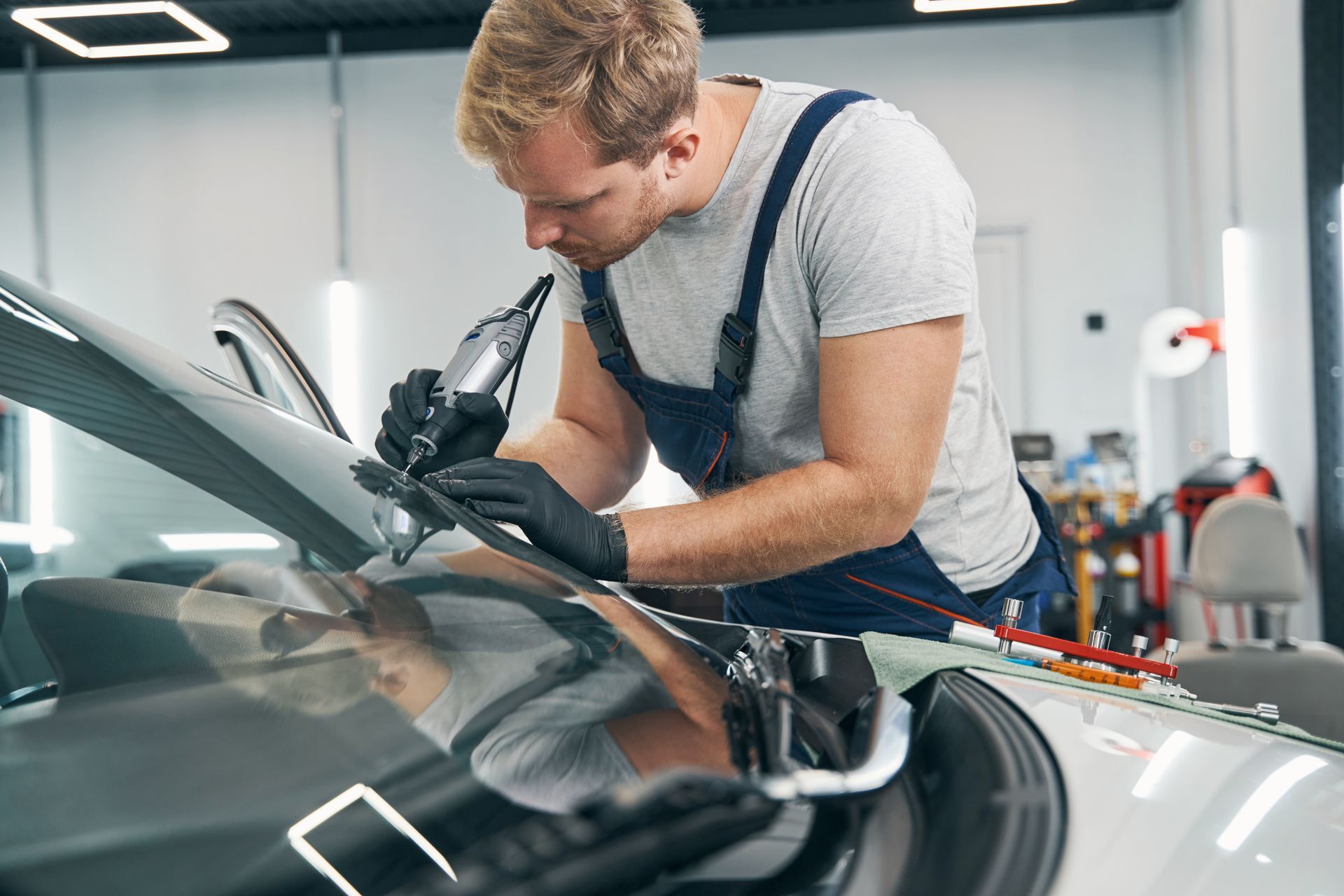A person in a gray shirt and blue overalls uses a handheld power tool to repair a crack on a car windshield in a garage.
