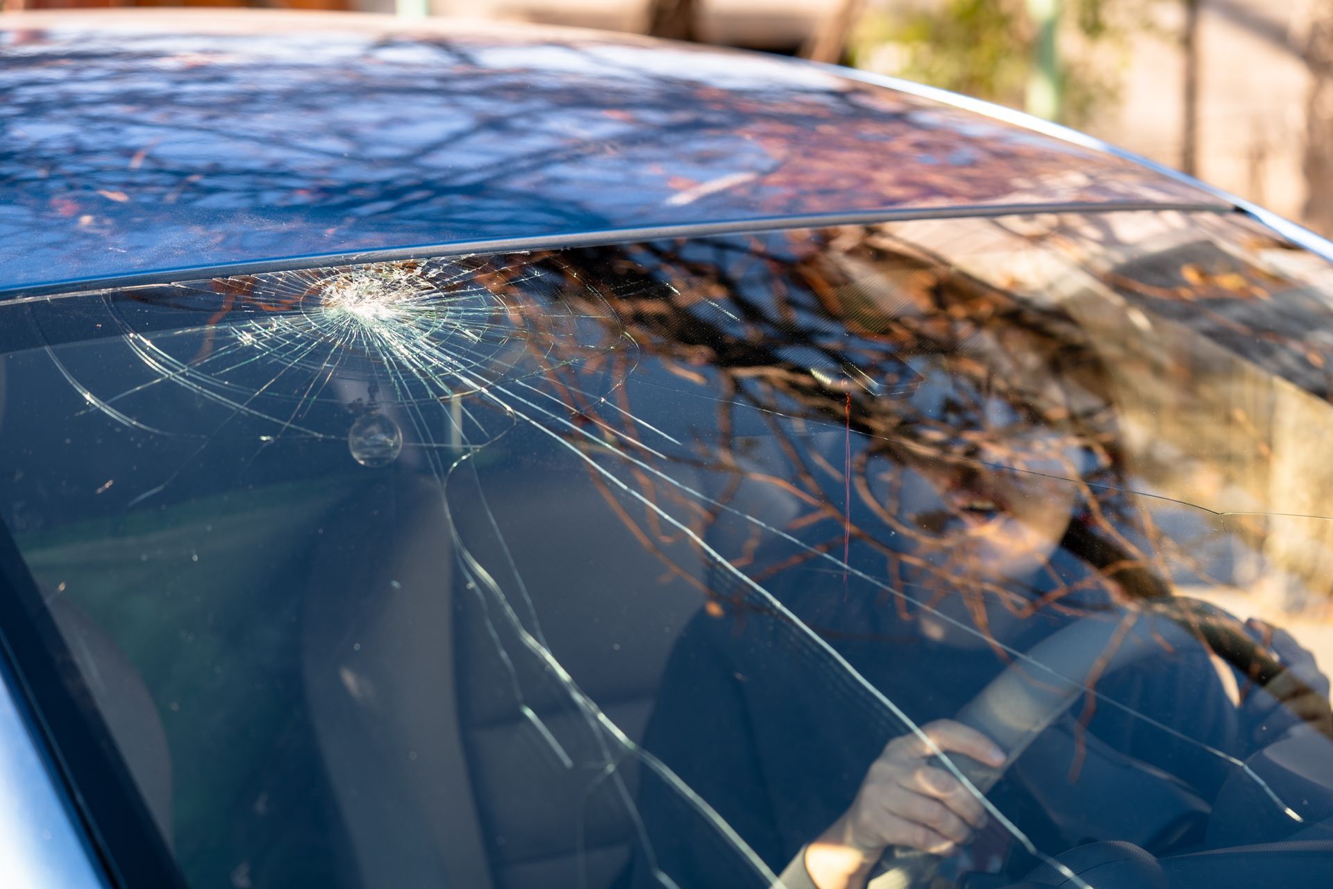 A cracked windshield on a blue car obscures a person driving; the sunlight gleams on the broken glass.