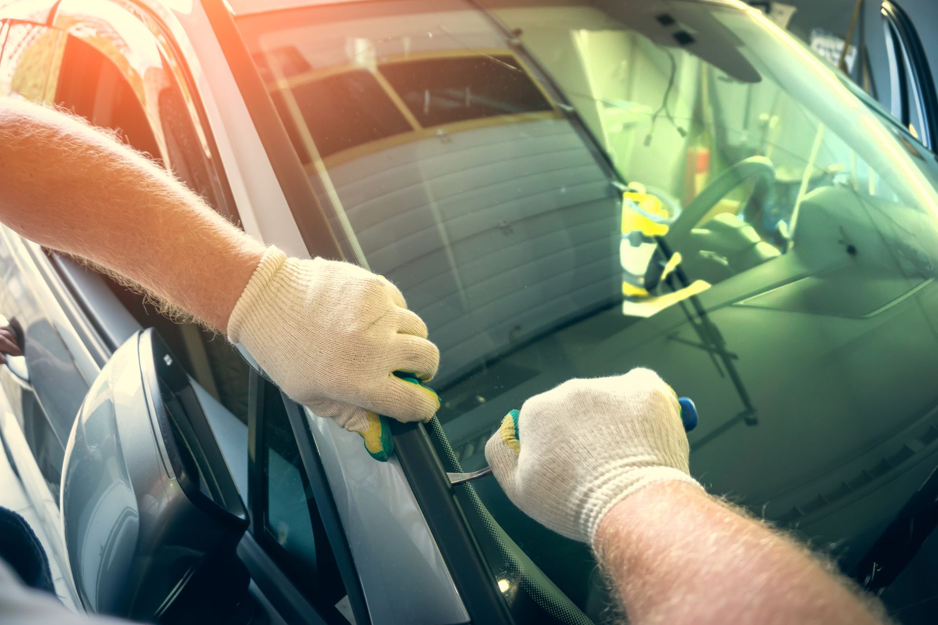 A worker wearing gloves uses a tool to remove a car's windshield seal in a garage.