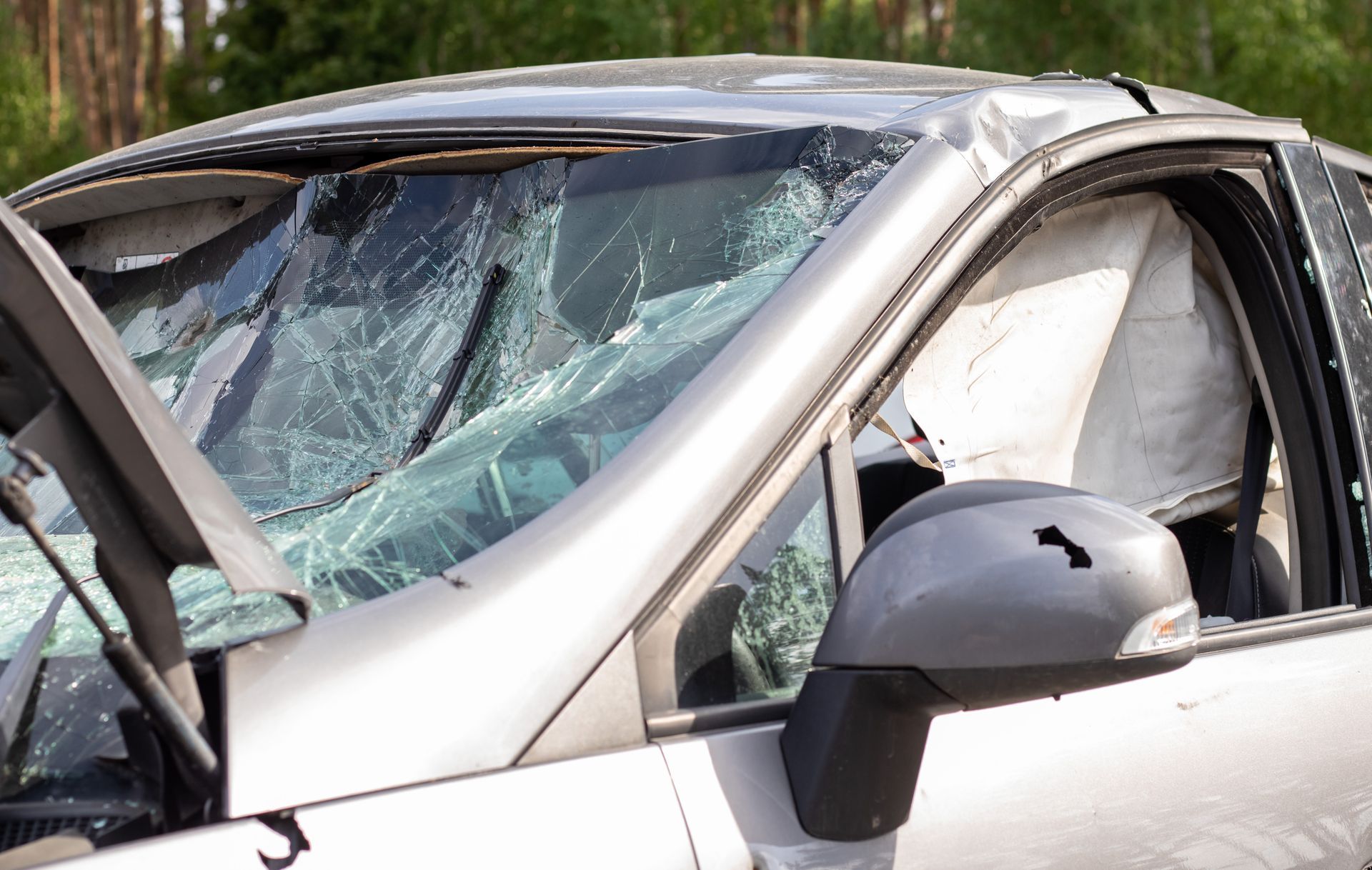 A silver car with a shattered windshield, crumpled hood, and deployed side-curtain airbag.