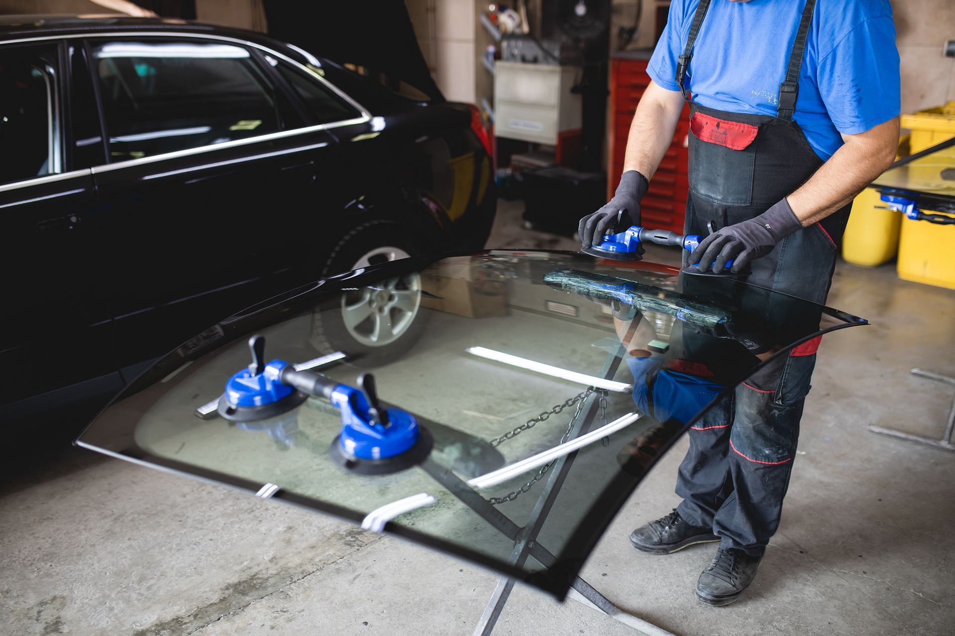 A mechanic handling a large car windshield using suction cup lifters in an auto repair shop.