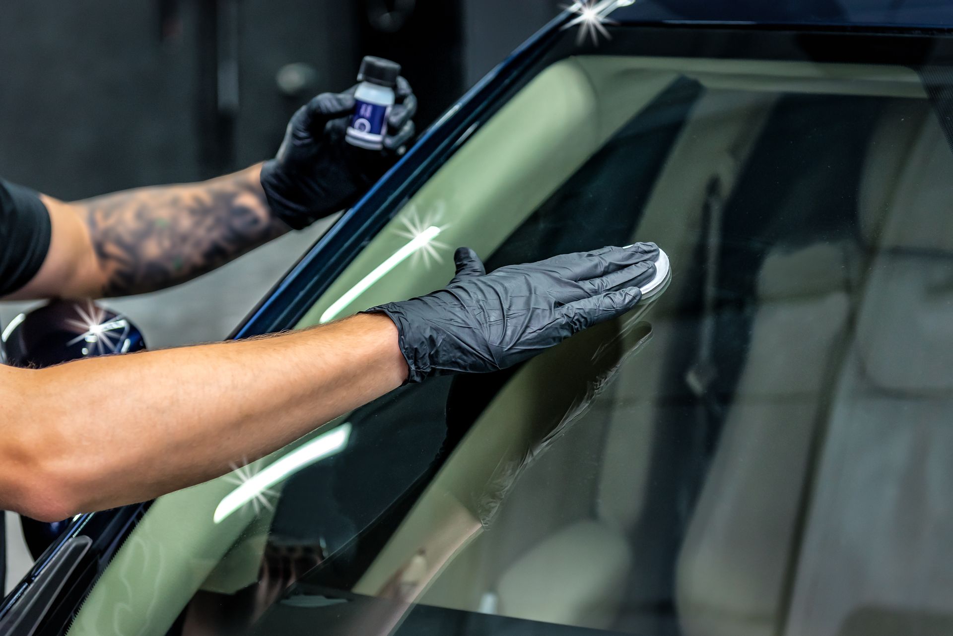 A person wearing black gloves applies a glass coating product to a car windshield in an indoor garage.
