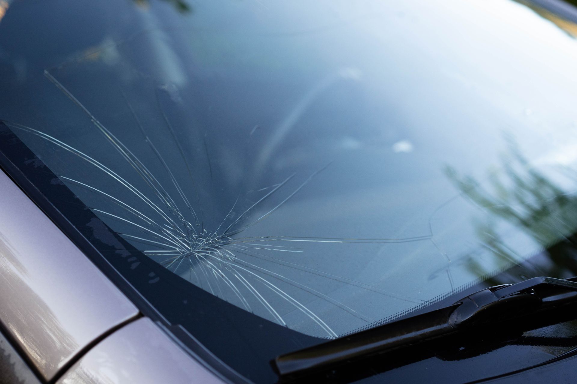 A damaged car windshield featuring a prominent spiderweb crack pattern emanating from a central impact point.