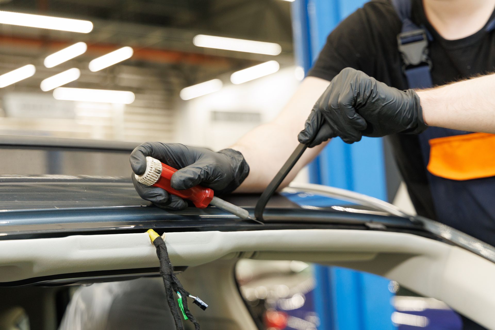 A technician in black gloves uses a pry tool and screwdriver to remove trim from a car’s roof in a garage.