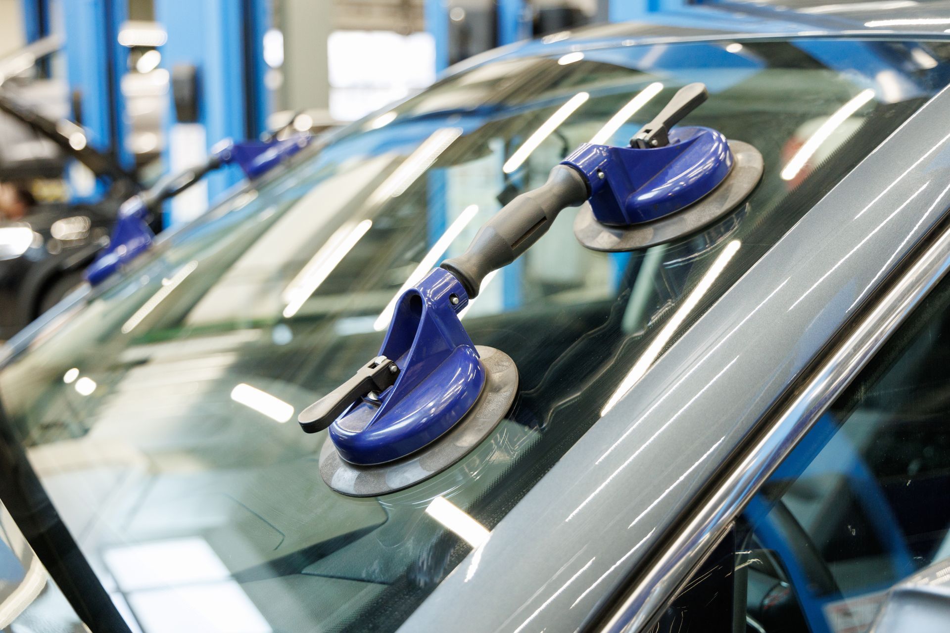 Two blue vacuum suction lifters attached to a car's windshield inside a repair shop.