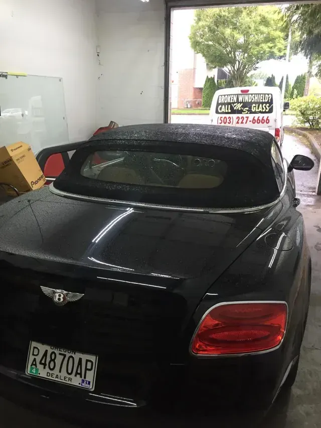 A dark Bentley convertible with a black soft top parked inside a garage, with a white service van visible outside.