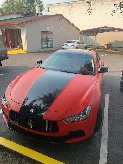 A matte red Maserati with a glossy black hood stripe parked in an outdoor lot near a building.