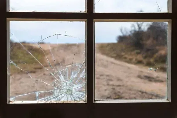 A pane of glass with a spiderweb crack pattern, looking out onto a dirt road and a rural landscape.