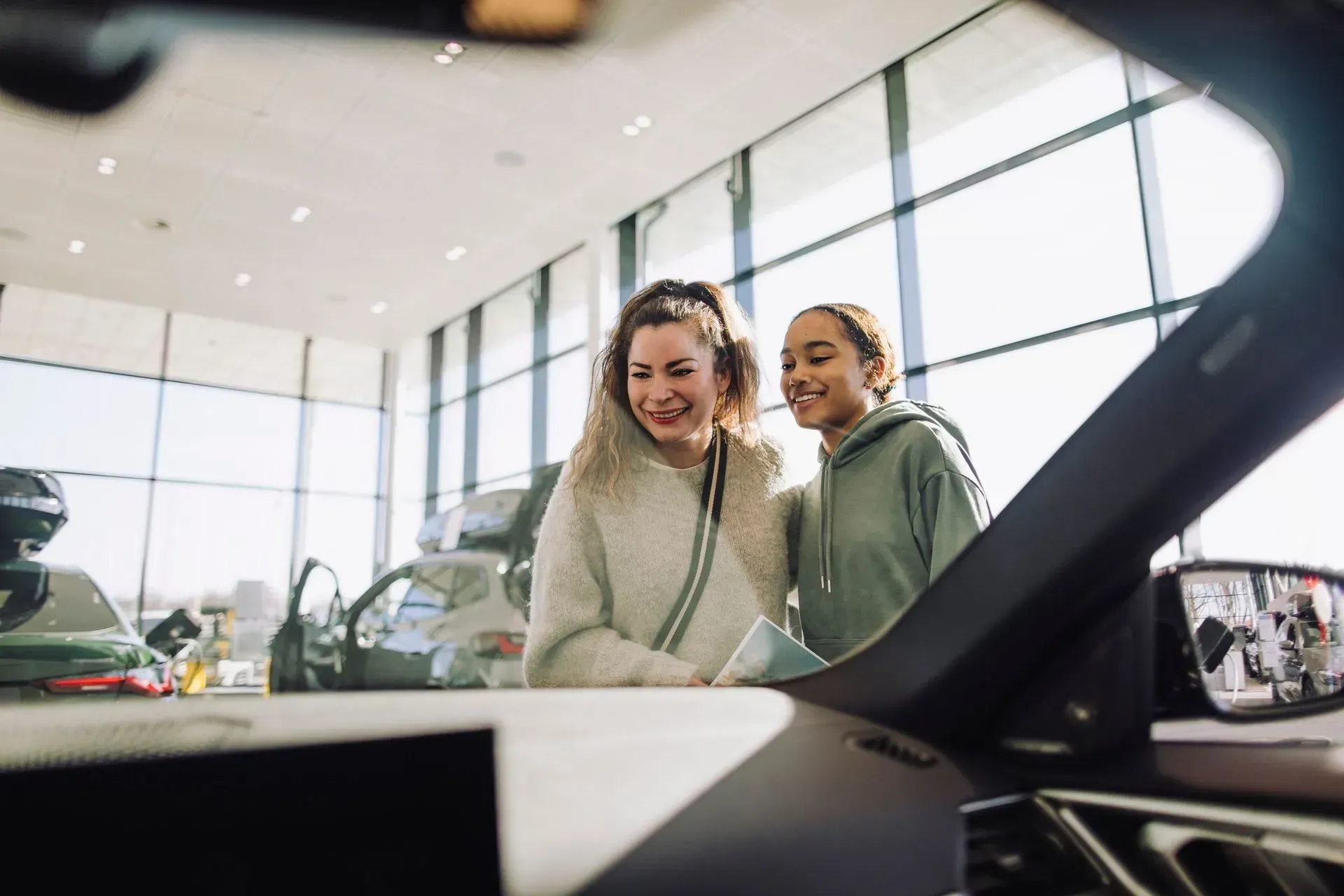 Two women smiling, looking at a car inside a dealership. Bright, natural lighting.