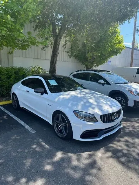 A white Mercedes-Benz coupe parked in a lot next to a white SUV under tree shade.
