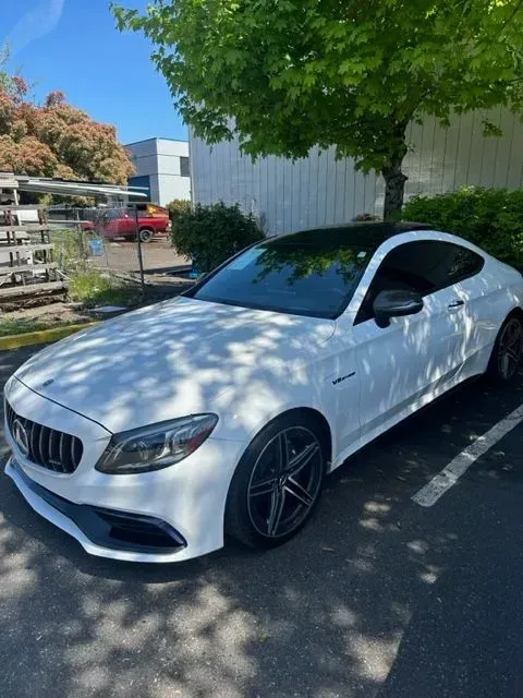 A white Mercedes-Benz C-Class coupe parked in a lot under a green tree on a sunny day.