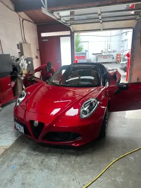 A bright red Alfa Romeo 4C parked inside an automotive workshop with a mechanic standing by the open driver’s side door.