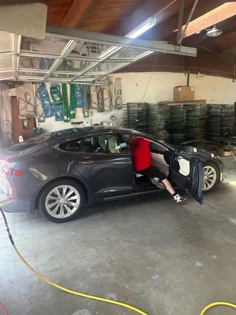 A person in a red shirt is sitting in the driver's seat of a dark grey sedan inside a garage with storage racks.