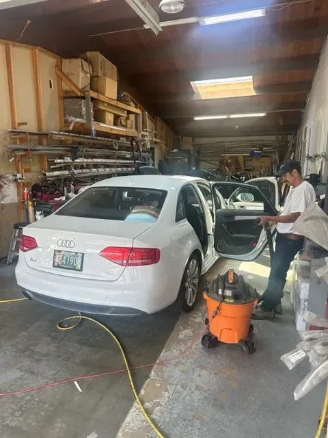 A person in a garage vacuums the interior of a white Audi sedan with its doors open.