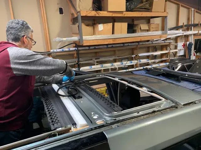 A person wearing a maroon and grey top and blue gloves works on the open sunroof mechanism of a vehicle in a workshop.