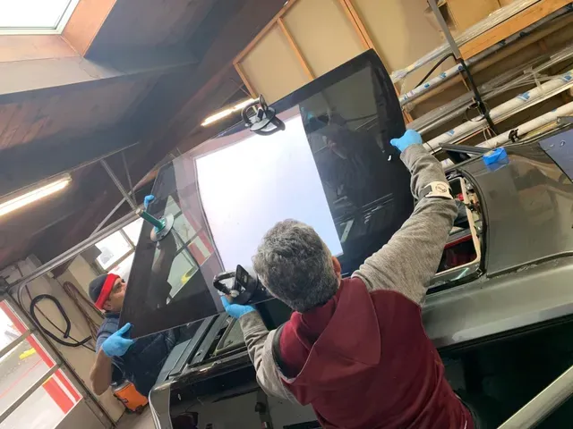 Two technicians in blue gloves install a new windshield onto a vehicle inside a bright automotive workshop.