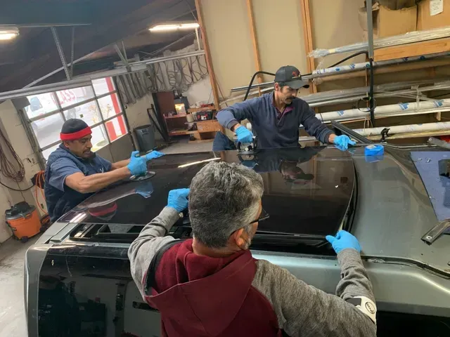 Three people in blue gloves work together to install a large glass sunroof panel onto the roof of a vehicle in a shop.