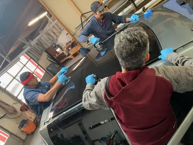 Three technicians wearing blue gloves work together to install a large glass panel onto a vehicle roof in a workshop.