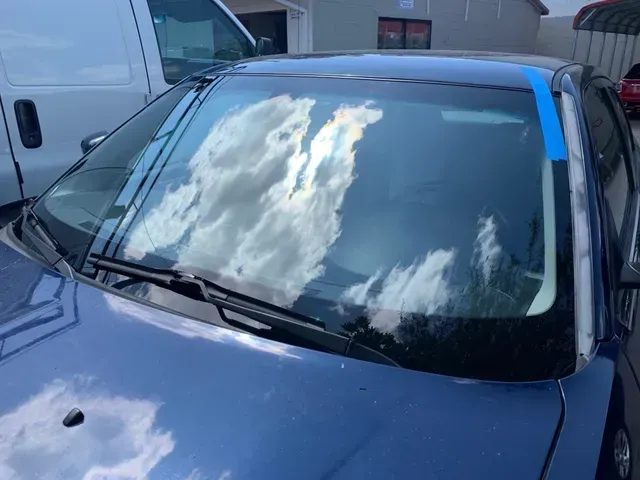 A blue car parked outdoors, featuring a replaced windshield held in place by blue painter's tape along the A-pillar.