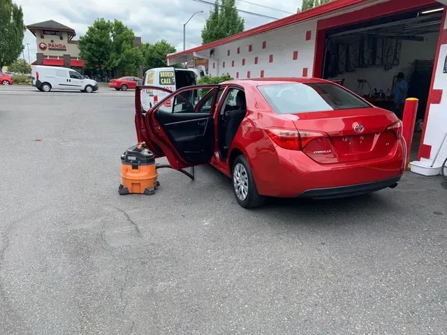A red Toyota sedan parked at a car wash with its doors open and a shop vacuum on the pavement nearby.