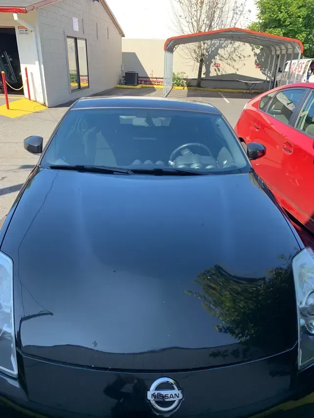 A black Nissan 350Z parked in a sunny outdoor lot with a red car to its right and a building in the background.