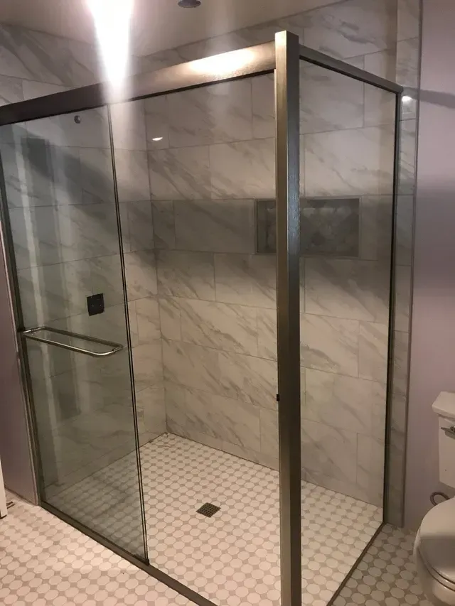 A walk-in shower with marble-style wall tiles, a glass door with a silver handle, and white hexagonal floor tiles.