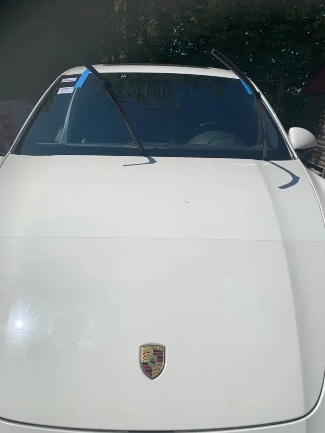 A front-facing white Porsche with its windshield wipers raised, parked outdoors on a sunny day.