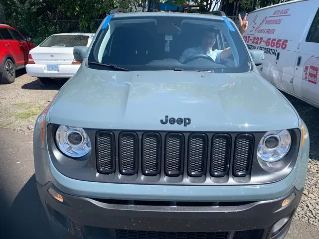 A light gray Jeep Renegade parked outdoors, with a driver in the front seat waving at the camera.