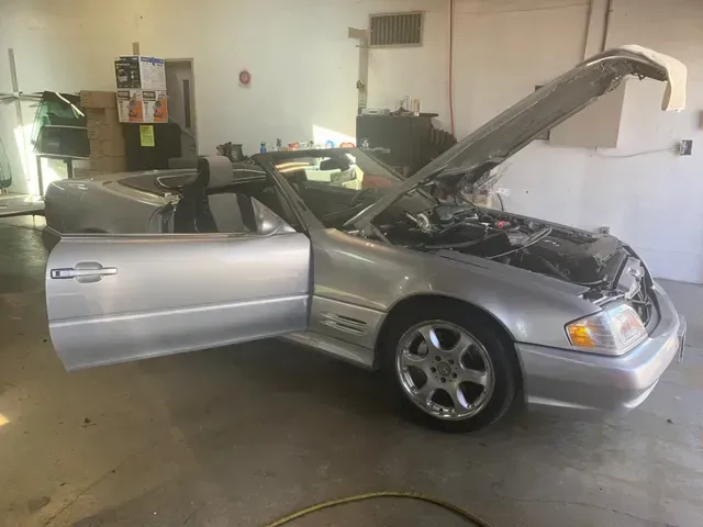 Silver convertible Mercedes-Benz SL-class with its hood open and driver-side door ajar inside a garage.